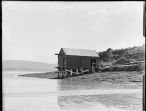 Two young men [Edgar and Owen Williams?] in striped vests standing in front of a boatshed on a rocky beach of unknown location with forest covered hills beyond, Catlins District, South Otago Region