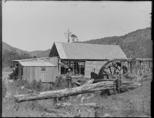 Two unidentified boys holding a rifle and rabbit in front of a derelict sawmill with a waterwheel, Catlins District, South Otago Region