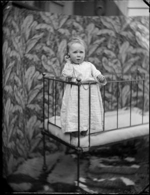 Portrait of a young Edgar Williams in nightgown standing in a cot holding a pocket watch at the family house, 'View Bank', Maitland Street, Dunedin