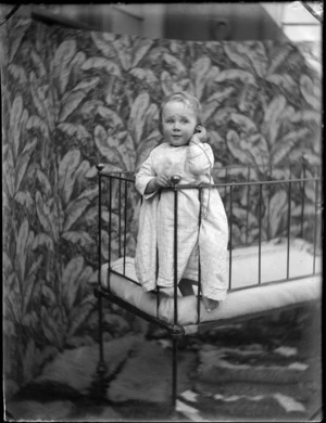 Portrait of a young Edgar Williams in nightgown, standing in a cot holding a pocket watch at the family house, 'View Bank', Maitland Street, Dunedin