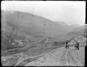 Two unidentified men with bicycles on [Porters Pass?] with tussock covered hills and the West Coast Road to Arthur's Pass, Canterbury Region