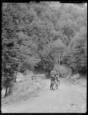 William Williams with a bicycle on a hill section of the [West Coast Road?] to Arthur's Pass surrounded by sub-alpine forest, Canterbury Region