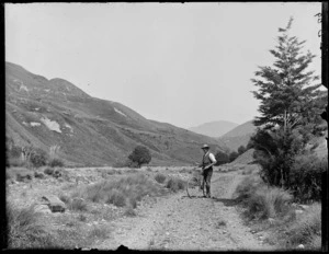 Owen Williams and bicycle on the [West Coast Road?] from [Porters Pass?] with tussock covered hills, Selwyn District, Canterbury Region