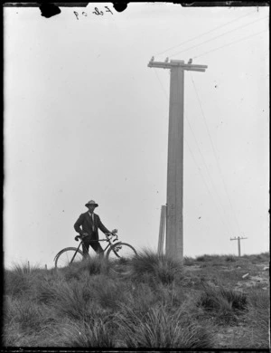 Owen Williams and bicycle on a tussock covered hill next to a power pole, Selwyn District, Canterbury Region