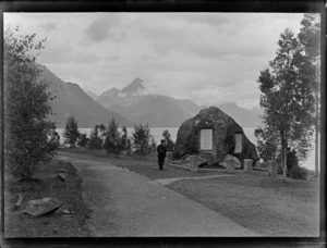 An unidentified man by the Robert Scott memorial boulder erected in 1913 within Queenstown Peninsula Gardens, with Lake Wakatipu and Walter Peak beyond, Central Otago Region