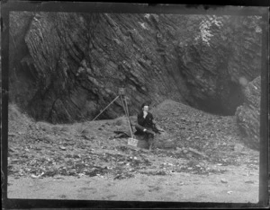 An unidentified photographer with camera and tripod sitting in front of a rock wall at an unknown beach eating a sandwich, [Dunedin Region?]