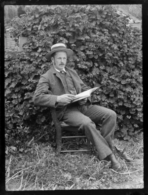Portrait of Thomas 'Tom' H Wyatt sitting in a chair reading a book at an unknown backyard location, [Dunedin Region?]