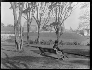 Lydia Williams seated on a park bench, looking towards the glasshouse, Auckland Domain