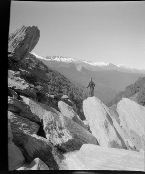 Part one of a stereograph showing unidentified mountaineer with ice axe standing on a rock, with glacier and mountain peaks beyond, West Coast Region