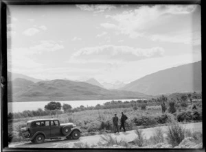 Two unidentified men and touring car on a shingle road beside Lake Wanaka looking to the Matukituki Valley and [Mount Aspiring?], Central Otago Region