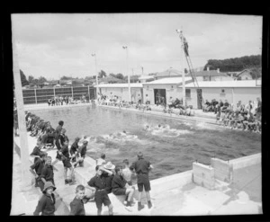 A large group of school children at a public swimming pool watching a boy's swimming race