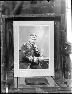 Brass band leader, with large handlebar moustache wearing band uniform, holding a conductors baton, sitting next to a trumpet on table