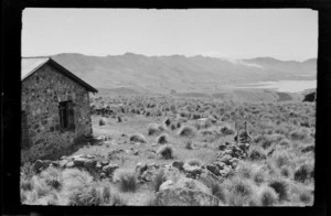 Landscape view of valley floor, lake and mountains, including stone house and wall, probably Banks Peninsula