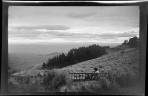 An unidentified woman sitting on a Port Hills walking track looking out over a tussock slope and a pine tree grove to the Canterbury Plains beyond, Banks Peninsula, Canterbury Region