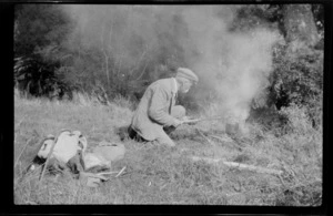 William Williams boiling a billy in bush area, walking canes and back packs in the foreground, Stewart Island, (Rakiura)