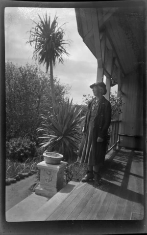 Unidentified woman on a veranda of house with flax bush and flowers in garden, Stewart Island (Rakiura)