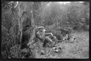 William Williams drinking from a cup while leaning on a tree, in the bush, Stewart Island (Rakiura)