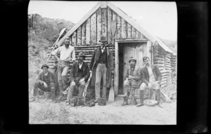 Six unidentified men outside a hut in bush, location unidentified
