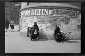 Two female street vendors, one selling shoelaces, sitting in front of a wall pasted with advertising posters, Marseille, France