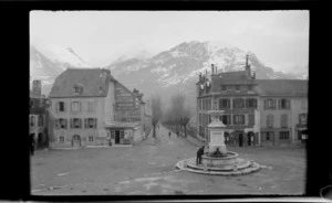 Village square, with fountain, billboard advertising Olibet biscuits and Byrrh, hotel on corner next to bridge, snowy mountain beyond, Laruns, Pyrenees-Atlantiques, France