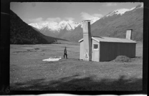 Man, probably B N Teague, folding a tent in front of alpine hut with view of mountains, during attempt on Stargazer, Fiordland, Otago region