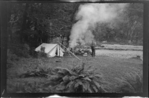 Man, probably B N Teague, at his campsite, with tent and campfire on a valley floor, during attempt on Stargazer, Fiordland, Otago region