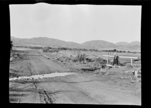 A road with hills in background, including a woman [Lydia Williams?] standing on a small footbridge over a stream, Hamner Springs, Hurunui district, Canterbury region