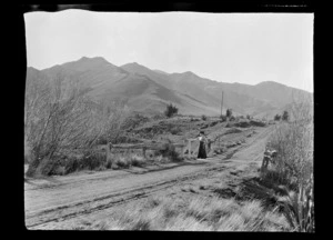 A road, with hills in background, and a woman [Lydia Williams?] standing next to a gate, Hamner Springs, Hurunui district, Canterbury region