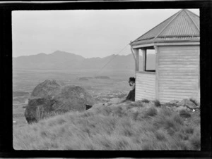 [Lydia Williams] with binoculars sitting next to wooden lookout, looking down over large rock and farmland below, Hanmer Springs forest plantation, Canterbury Region