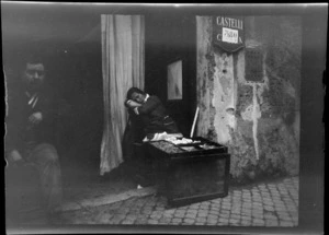 Unidentified street vendor resting with head on arms, beside a stone wall with a sign reading 'Castelli 380', Barcelona, Spain