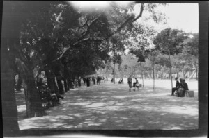 People walking and sitting on seats in tree-lined promenade, including a child with a scooter, Naples, Italy