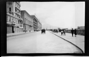 Pedestrians on walkway near sea wall, including a horse and buggy on road, with buildings on the left and Mount Vesuvius in the distance, Naples, Italy