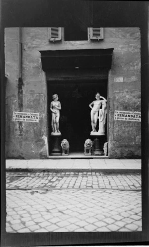 Statues, including a female, male and two lions in a shop door, with the number '160' and 'La Cerimonia Al Colosseo E Rimandata A Giorno Da Destinarsi' written on the concrete wall, Rome, Italy