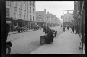 Street scene, people, horses and carts, vehicles, including woman street vendor sitting by side of road, Killarney, County Kerry, Ireland