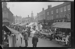 Street market, vendors selling goods and buyers on street, including shop buildings in village square, [Manchester], England