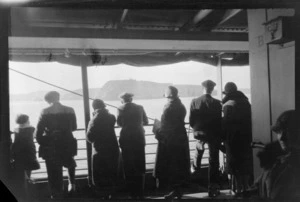 Scene on the passenger deck of a ship, showing a group of unidentified men, women and children looking to an unidentified coastline