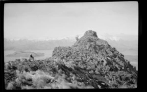 Rocky hilltop with an unidentified woman sitting amongst tussock, snow-capped peaks bordering harbour below, [Heathcote Valley, Canterbury region?]