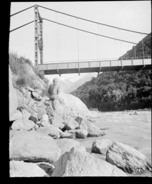 An unidentified man sitting on boulders next to a river, beneath a suspension bridge, [Fox Glacier, West Coast Region?]