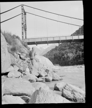 An unidentified man sitting on a boulder next to river, beneath a suspension bridge, [Fox Glacier, West Coast Region?]