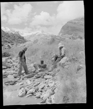 Two unidentified men lighting a campfire next to a river, Fox Glacier, West Coast Region