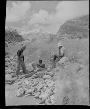 Two unidentified men lighting a campfire next to a river, Fox Glacier, West Coast Region