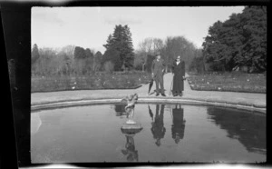 William and Lydia Williams standing by edge of round pond with cherub in centre, with flower beds and trees in public garden area, unknown location, England