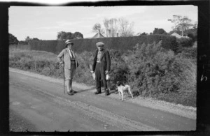 An unidentified man and woman with a dog, standing on a country road, the man holds dead rabbits, location unidentified