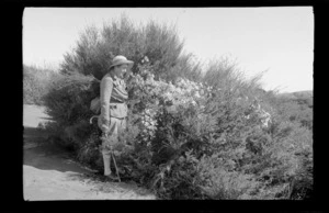 Alice Williams dressed in walking attire, standing next to a flowering vine, [Kawhia, Waikato Region?]