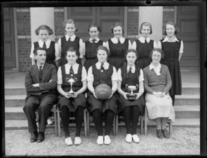 Girls' netball team and two unidentified teachers, with cup trophies, outside a brick building, at Westport Technical High School, West Coast Region