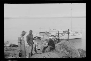 Edgar Williams, Owen Williams and an unidentified fisherman, showing Owen holding a catch of fish, on rocky shore next to small dock and boat, probably Waikato Region