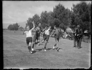Group of unidentified schoolboys crossing the finish line during a running race, officiated by two men, spectators sit on chairs alongside the track, at Westport Technical High School, West Coast Region
