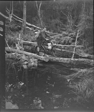 Alister McLellan wheeling a bicycle acoss a log bridge in the bush, probably West Coast Region