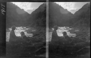 A unidentified man smoking a pipe, sitting with a dog next to river, probably West Coast Region