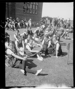 Group of unidentified girls sitting on grass at a school sports day, outside a large brick building, Westport Technical High School, West Coast Region